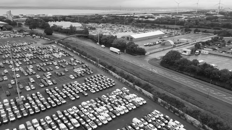 Rows of finished vehicles parked across a port storage compound, with terminal warehouses and the quayside beyond.
